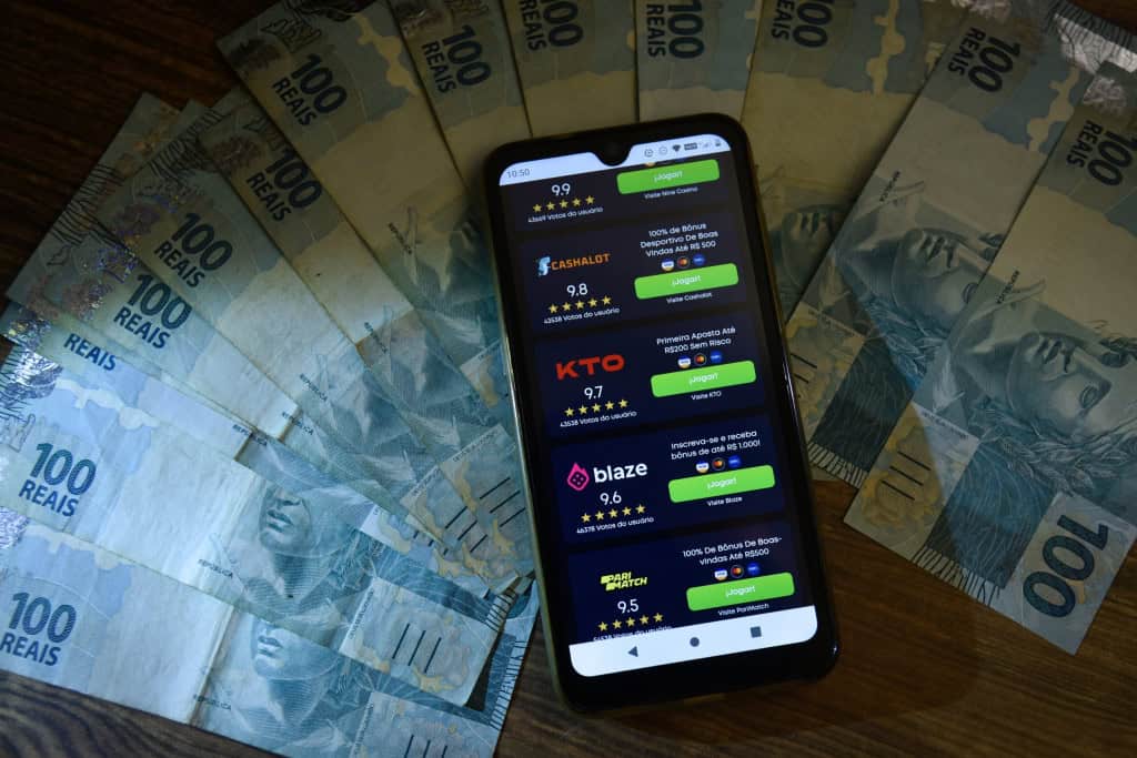 Várias notas de R$ 100 com um celular em cima, onde há o logotipo de várias bets