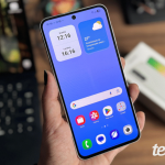 Imagem mostra mão segurando celular Samsung Galaxy A56 em primeiro plano, com fundo desfocado e caixa do aparelho ao fundo