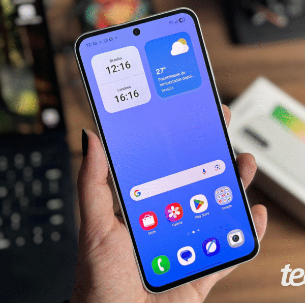 Imagem mostra mão segurando celular Samsung Galaxy A56 em primeiro plano, com fundo desfocado e caixa do aparelho ao fundo