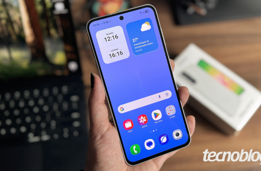 Imagem mostra mão segurando celular Samsung Galaxy A56 em primeiro plano, com fundo desfocado e caixa do aparelho ao fundo