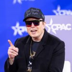 Elon Musk sentado, de óculos escuros, boné, falando e com o dedo indicador direito em riste