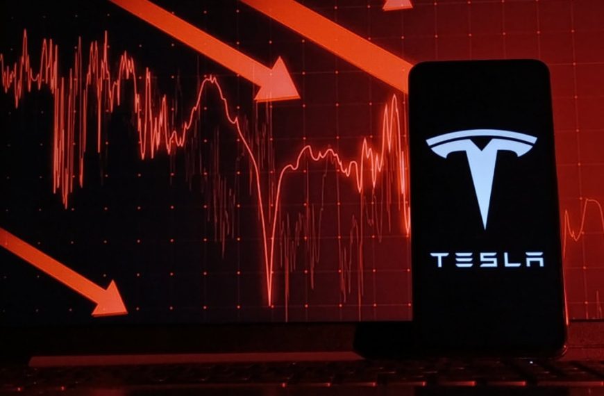 Logo da Tesla em um smartphone com indicadores financeiros atrás